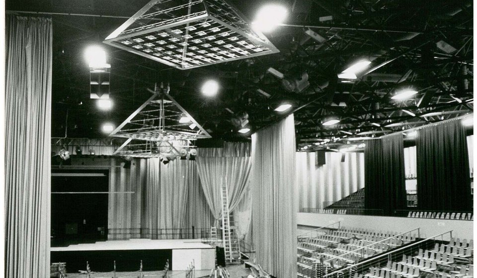 Von der Decke der Halle hängen pyramidenförmige Scheinwerfer. Zwischen den Vorhängen, die ebenfalls von der Decke hängen, sieht man die Bühne und die seitlichen Tribünen. 