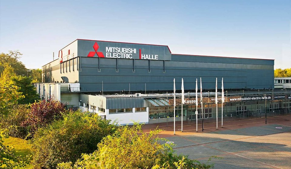 Mitsubishi Electric HALLE exterior view with car park in front. At the hall, banners with the words “Kasse” and “Eingang” are visible.