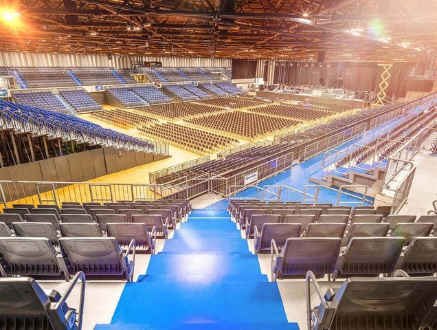 In der Mitte des Bildes sieht man einen blauen Teppich durch den Oberrang gehen. IM Innenraum sind mehr Stühle in Blöcken angeordnet. 