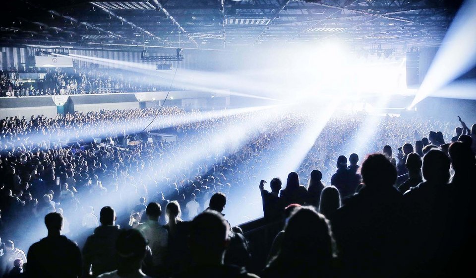 Blick aus dem Oberrang Richtung Bühne. Von der Bühne scheinen Lichtkegel durch den Innenraum. Die ganze Halle ist mit Menschen gefüllt. 