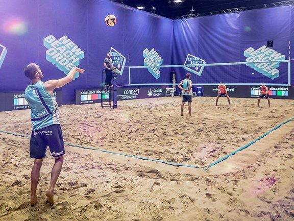 Beachvolleyballfeld mit Spielern. Im Vordergrund spielt ein Spieler den Ball auf die andere Seite des Netzes.