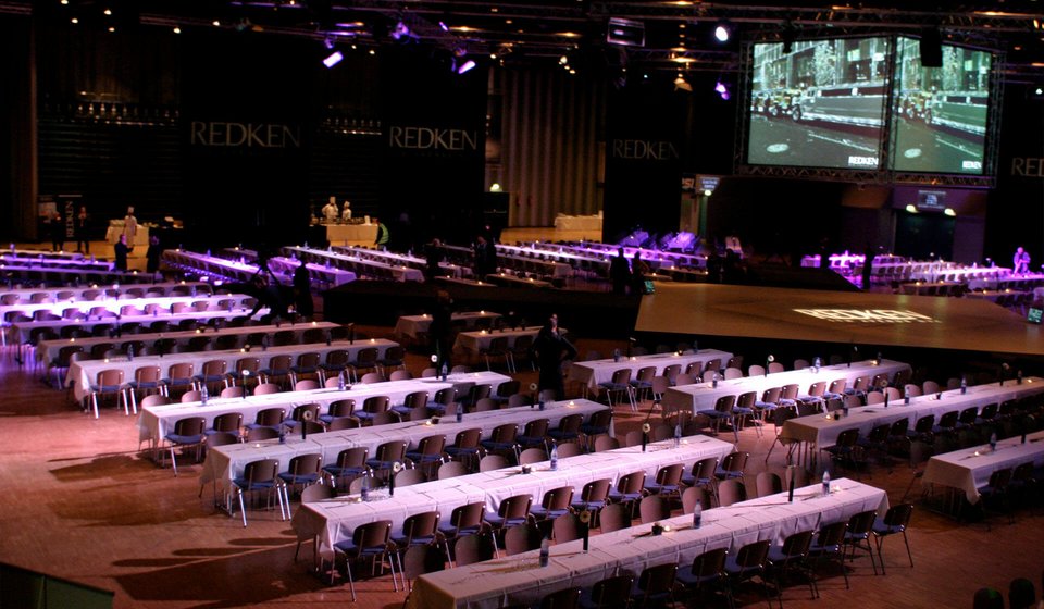 In der Mitte des Raum ist eine quadratische Bühne aufgebaut. Über der Bühne hängt ein Videowürfel. Rundherum sind längliche Tische mit Stühlen aufgebaut. 